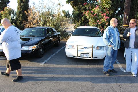 2003 Ford Crown Victoria - black and a white Ford sedan with brush guards sit parked while two men in denim talk beside them.