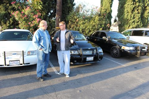 2003 Ford Crown Victoria - Two men converse between a white Ford sedan and black Ford sedans fitted with brush guards in a tree-lined lot.