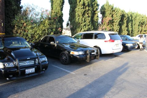 2003 Ford Crown Victoria - Two black Ford sedans, one with a brush guard, sit parked next to a white minivan along a tall green hedge.