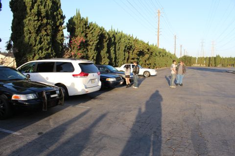 2003 Ford Crown Victoria - white minivan and dark sedans line a hedge while people stand in a wide-open paved lot in late afternoon light.