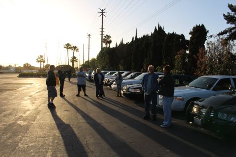 2003 Ford Crown Victoria - People gather in a parking lot beside a row of sedans, one with a green brush guard, under a low evening sun.