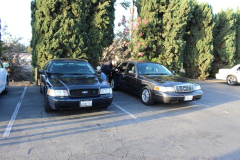 2003 Ford Crown Victoria - Two dark Ford Crown Victoria sedans park nose-to-tail in a lot as a person leans into one car's door.