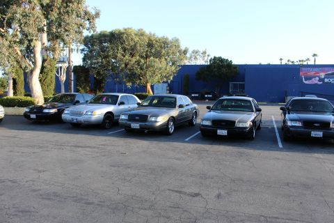 2003 Ford Crown Victoria - row of Ford sedans in silver, gold, and black sit parked in a lot in front of a long blue building.