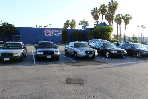 2003 Ford Crown Victoria - Black and silver Ford sedans park in a lot beside a blue building topped with signs and palm trees.