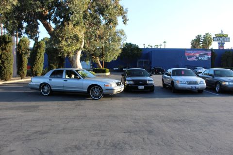 2003 Ford Crown Victoria - silver Ford sedan with chrome wheels leads a row of black and tan sedans parked near a blue building.