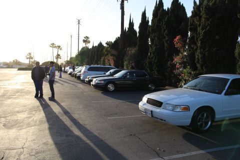 2003 Ford Crown Victoria - white Ford Crown Victoria sits in the foreground with a row of parked cars and a few people behind it at sunset.