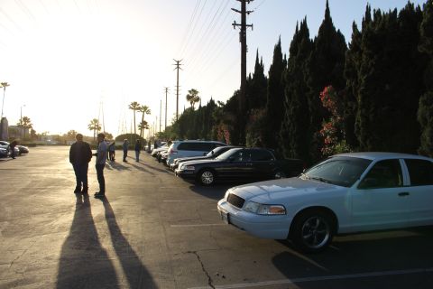 2003 Ford Crown Victoria - white Ford Crown Victoria and a row of dark sedans fill a parking lot as people stand casting long shadows in the low evening sun.