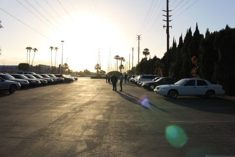 2003 Ford Crown Victoria - white Ford Crown Victoria sits at right while people stand in a paved lot lined with parked cars under a low backlit sun.