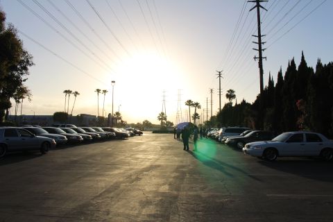 2003 Ford Crown Victoria - white sedan sits at right while rows of parked cars line a paved lot under a bright low sun and tall transmission towers.