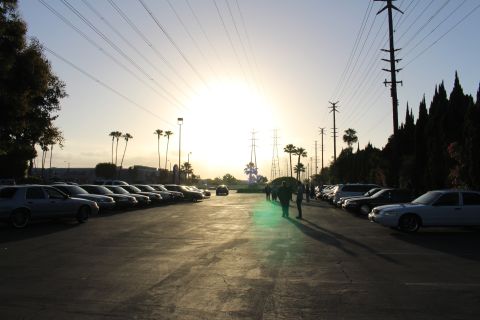 2003 Ford Crown Victoria - white sedan and rows of parked cars edge a paved lot, silhouetted figures and palm trees against a low bright sun.