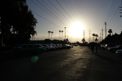 2003 Ford Crown Victoria - Parked cars line the left side of a dim paved lot as the sun sets behind palm trees and steel transmission towers.