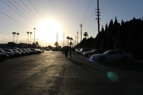 2003 Ford Crown Victoria - white Ford Crown Victoria sits at right while silhouetted cars and a walking figure fill a paved lot under a low sun.