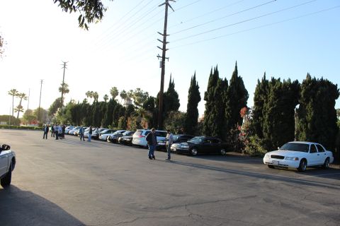 2003 Ford Crown Victoria - white Ford Crown Victoria sits at right as people stand among parked sedans in a bright paved lot beside cypress trees.