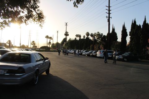 2003 Ford Crown Victoria - silver sedan with a visible plate sits foreground-left as people walk a paved lot of parked cars under a low sun.
