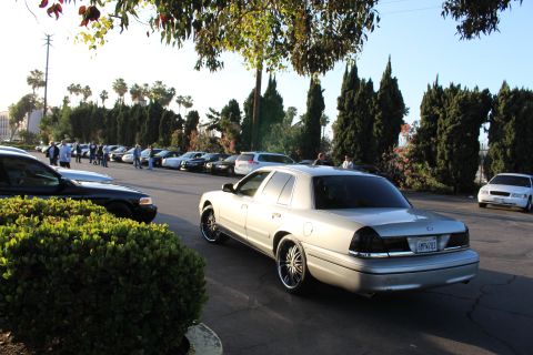 2003 Ford Crown Victoria - silver Ford Crown Victoria sits angled in a sunlit lot, its rear plate visible, with parked cars and people in the background.