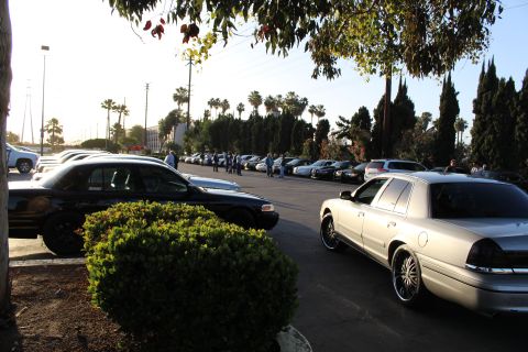 2003 Ford Crown Victoria - silver Ford Crown Victoria at right and a black sedan at left sit in a sunlit lot as people gather beyond rows of cars.