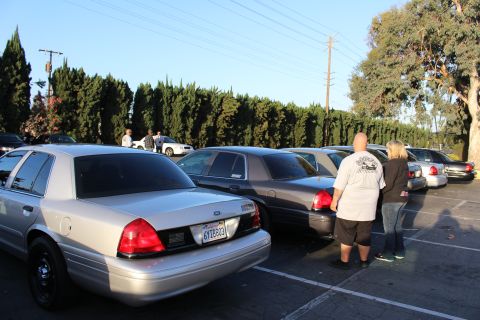 2003 Ford Crown Victoria - silver Ford Crown Victoria sits in the foreground as two people stand among parked sedans in a sunlit lot by cypress trees.