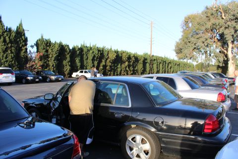 2003 Ford Crown Victoria - man leans into the open door of a black Ford Crown Victoria sedan parked in a lot bordered by tall cypress trees.