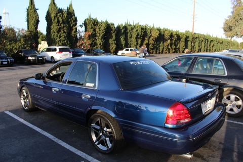 2003 Ford Crown Victoria - lowered dark blue Ford Crown Victoria sedan on chrome multi-spoke wheels, parked in a tree-lined lot.