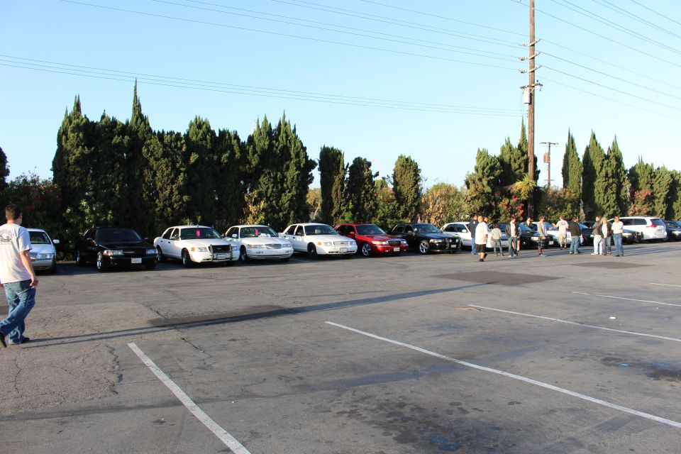 Row of parked sedans with people walking in a large lot, a person in white tee at left edge
