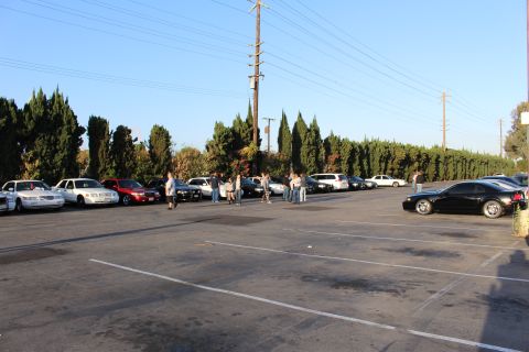 2003 Ford Crown Victoria - group of people stand and talk in a large lot between rows of parked sedans, with a black coupe parked at right.