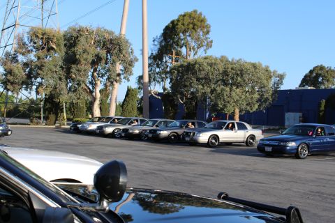 2003 Ford Crown Victoria - row of full-size sedans parked near a transmission tower, a dark blue car at right and a glossy black hood in front.
