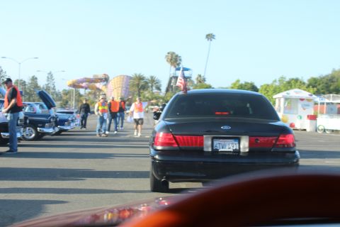 2003 Ford Crown Victoria - black Ford sedan drives through a sunny parking lot car show, with an American flag and other cars and people lining the lot.