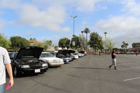 2003 Ford Crown Victoria - Several sedans parked with hoods open at a car meet under a cloudy sky as a man walks across the open parking lot.