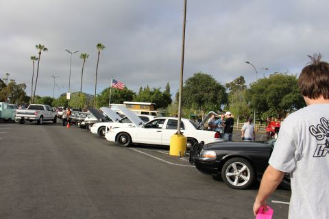 2003 Ford Crown Victoria - White and black sedans with raised hoods line a parking lot car meet near an American flag, a man in a gray shirt watching.