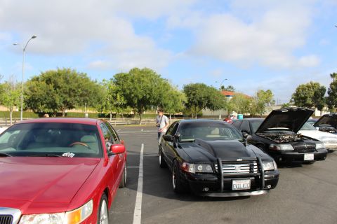 2003 Ford Crown Victoria - red sedan parked beside a black sedan fitted with a front push bar at a parking lot car meet under blue sky.
