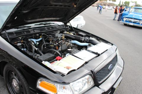 2003 Ford Crown Victoria - open engine bay of a black Ford at a car meet, with a judging sheet laid on the radiator support and other cars in the background.