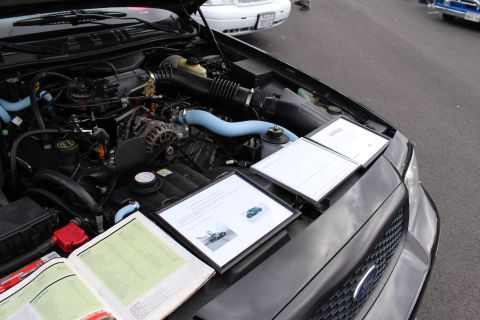 2003 Ford Crown Victoria - Ford engine bay with blue silicone hoses and several printed judging sheets propped on the fender at a car meet.