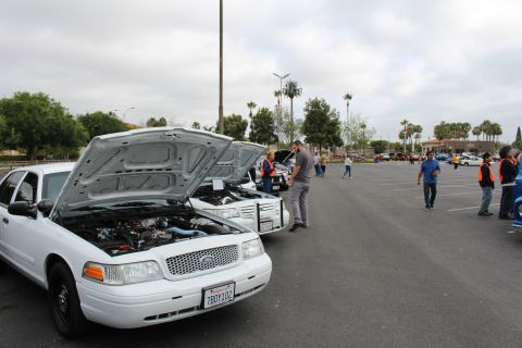 2003 Ford Crown Victoria - white sedan sits with its hood raised as people mill around a wide parking lot car meet under an overcast sky.