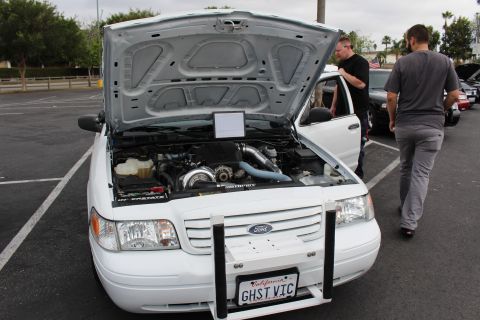 2003 Ford Crown Victoria - white Ford displays a turbocharged engine under its raised hood, personalized plate GHST VIC, as two men stand beside it at a meet.