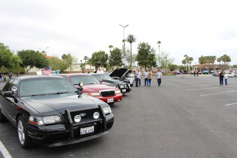 2003 Ford Crown Victoria - black sedan with a front push bar leads a row of parked cars at a car meet, an American flag flying and people gathered down the line.