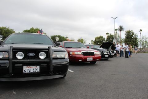 2003 Ford Crown Victoria - black Ford fitted with a push bar and spotlights leads a line of sedans at a car meet, a red Mercury behind and people gathered ahead.