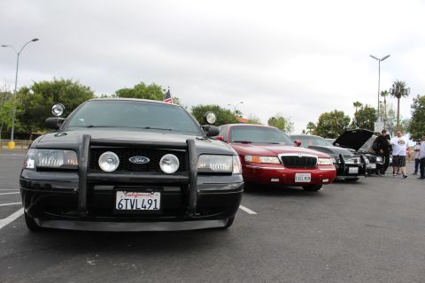 2003 Ford Crown Victoria - Front three-quarter view of a black Ford with push bar and spotlights, plate 6TVL491, heading a row of cars at an overcast car meet.