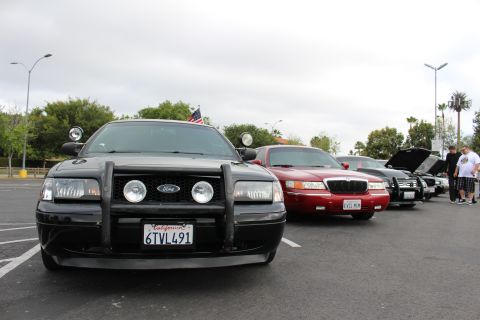 2003 Ford Crown Victoria - black Ford with push bar and spotlights and a red Mercury head a row of cars at a parking lot meet under a gray sky.
