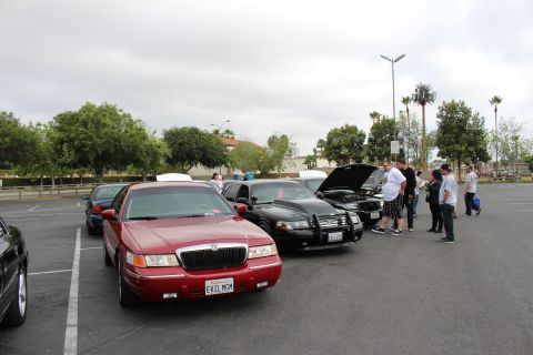 2003 Ford Crown Victoria - red Mercury sedan parks beside a black sedan as a group of people gather around open-hood cars at a parking lot meet.