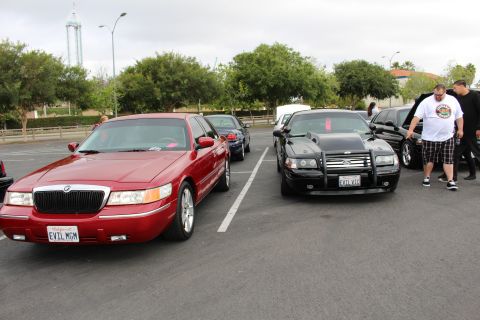 2003 Ford Crown Victoria - red Mercury parks beside a black sedan with a push bar and plate EVIL VIC at a car meet, men standing to the right.
