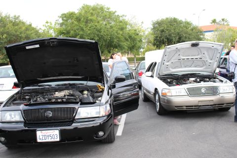 2003 Ford Crown Victoria - black sedan (plate 4ZDY503) and a gold Mercury sedan parked with hoods open at a car meet as people stand alongside.
