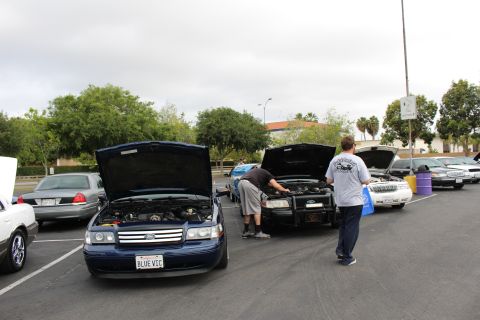 2003 Ford Crown Victoria - blue Ford sedan with vanity plate BLUE VIC and a black push-bar-equipped car sit with hoods raised as men work on engines at a meet.