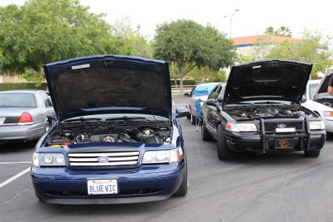 2003 Ford Crown Victoria - blue Ford Crown Victoria (plate BLUE VIC) beside a black Ford sedan with a brush guard, both with hoods raised in a parking lot.