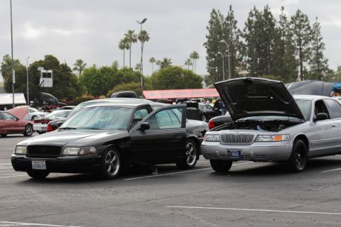 2003 Ford Crown Victoria - black Mercury full-size sedan parked beside a silver Mercury with its hood raised at a busy outdoor car meet with palm trees behind.