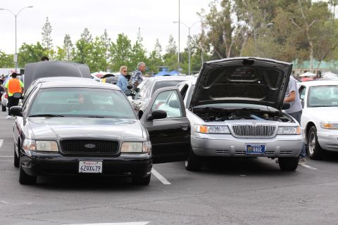 2003 Ford Crown Victoria - black Ford sedan (plate 6ZQX479) beside a silver Mercury with its hood raised and vanity plate RAGE at an outdoor car gathering.