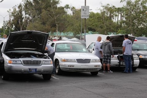 2003 Ford Crown Victoria - silver Mercury with vanity plate RAGE and a white Lincoln sedan parked with hoods raised as people gather around at a car meet.
