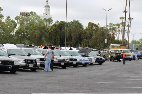 2003 Ford Crown Victoria - row of full-size sedans with hoods up parked beside a green vintage military-style vehicle at a car meet, clock tower behind the trees.