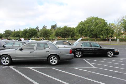 2003 Ford Crown Victoria - gray and a black Ford Crown Victoria sedan sit nose-to-tail in marked parking spaces with trees behind.
