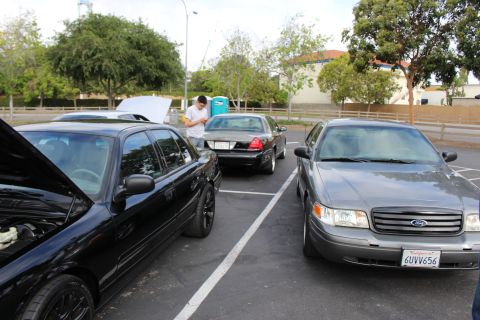 2003 Ford Crown Victoria - gray Ford sedan with California plate 6UVV656 parks beside black sedans, one with its hood up, at a car meet.