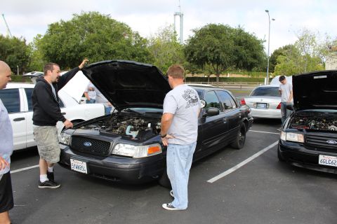 2003 Ford Crown Victoria - Several men gather around a black Ford Crown Victoria with its hood raised in a tree-lined parking lot.